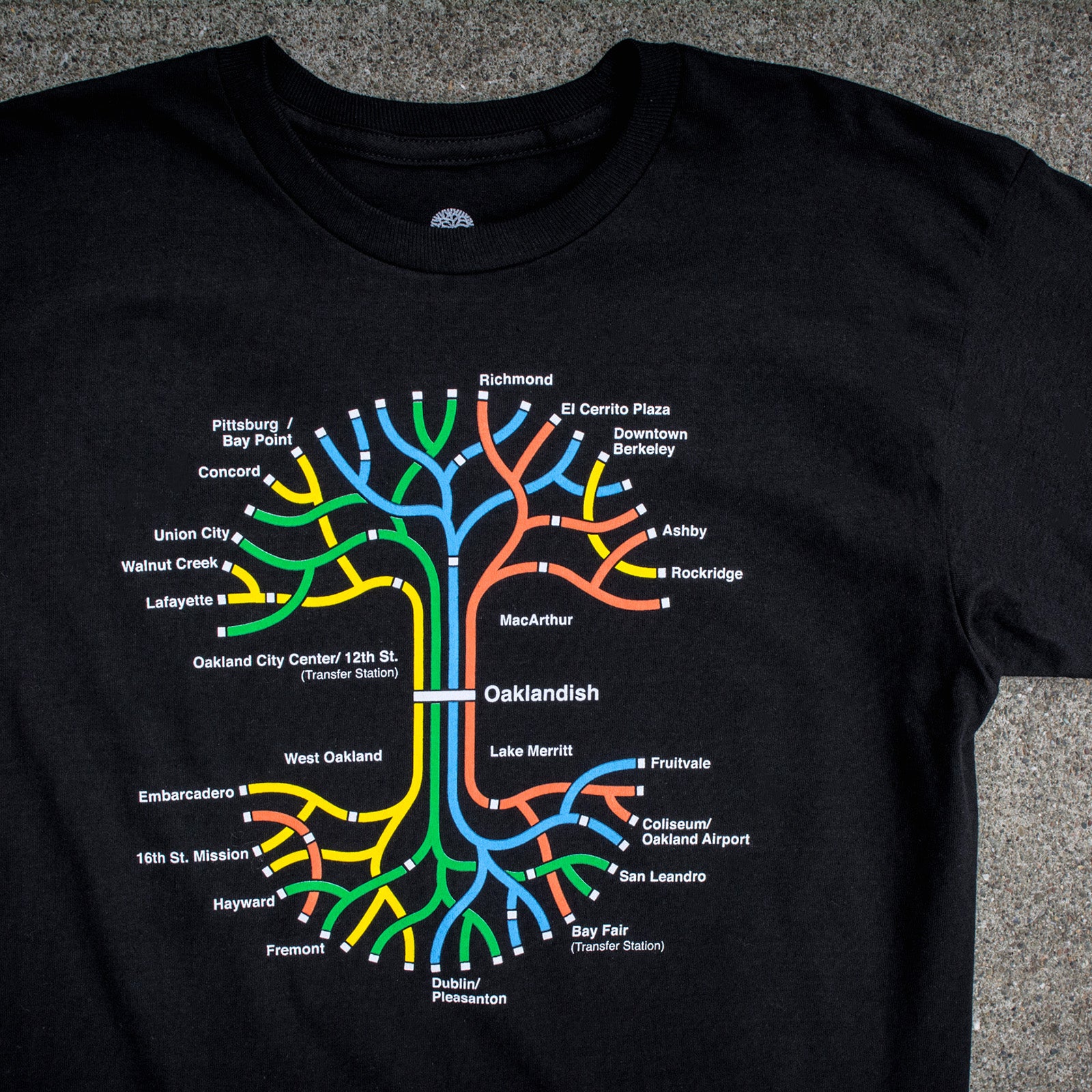 Close up a full-color BART transit map in the shape of the Oaklandish tree logo on a black t-shirt laying outdoors on cement.