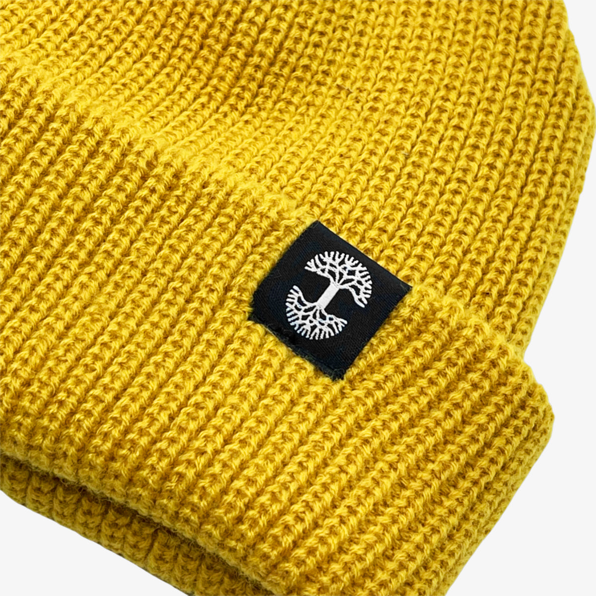 Close up of black and white Oaklandish tree logo tag on the left wear side of a shallow fit mustard yellow cuffed beanie.