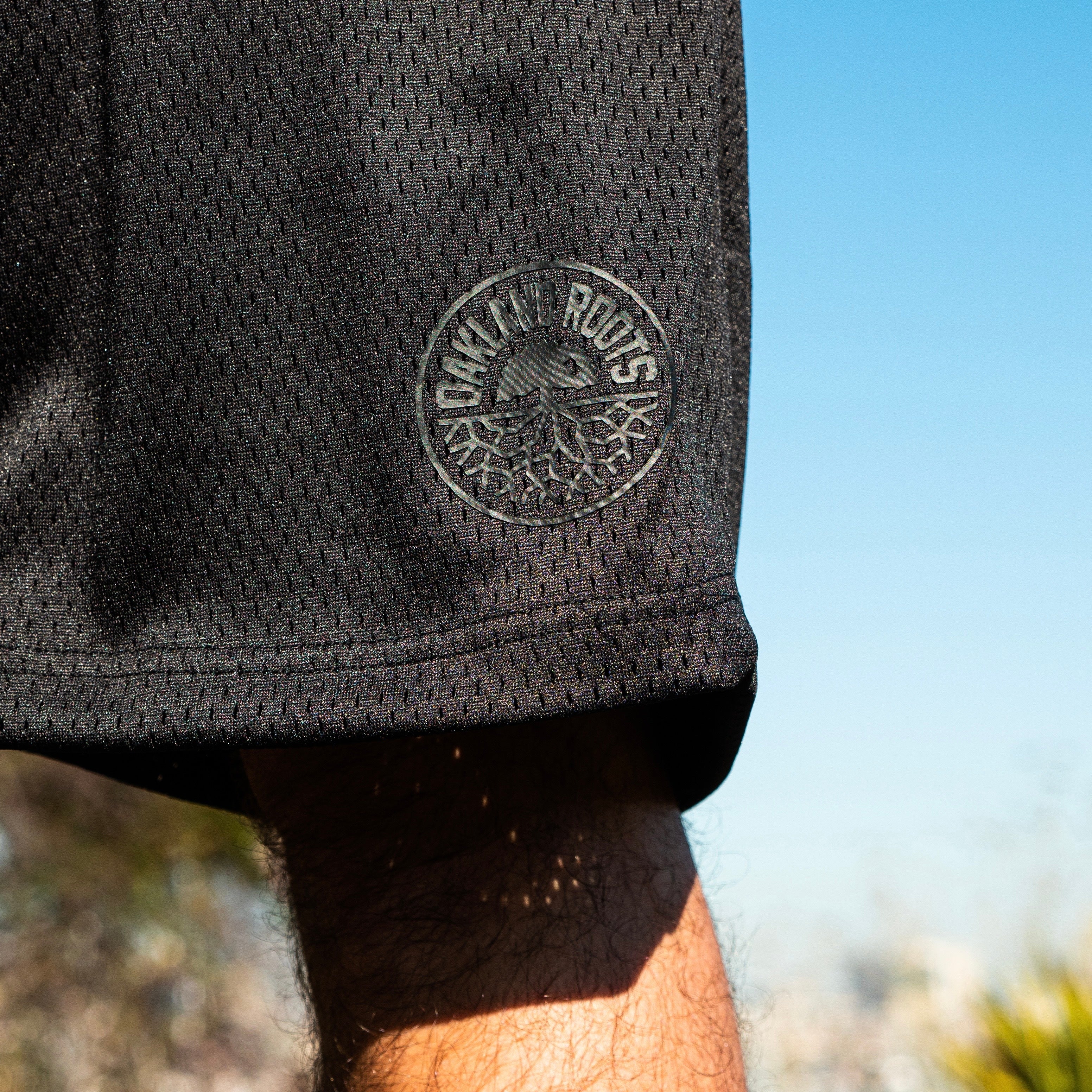 Close up of round Oakland Roots logo on shorts on a man’s leg outdoors with blue sky background.