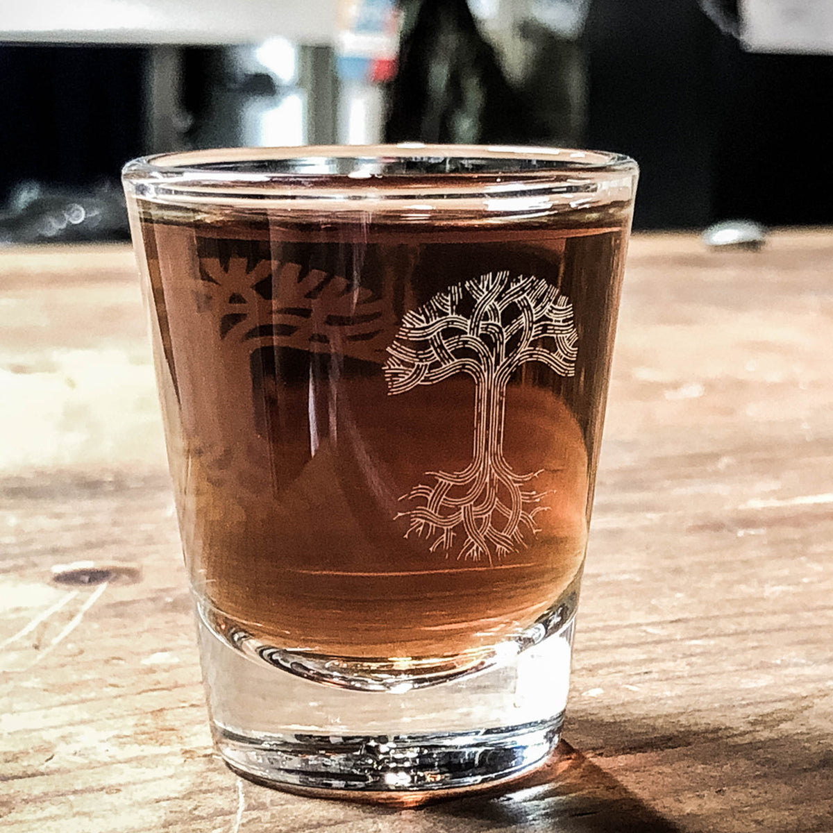 Clear glass shot glass with translucent Oaklandish tree logo full of brown liquor on a wood picnic table.