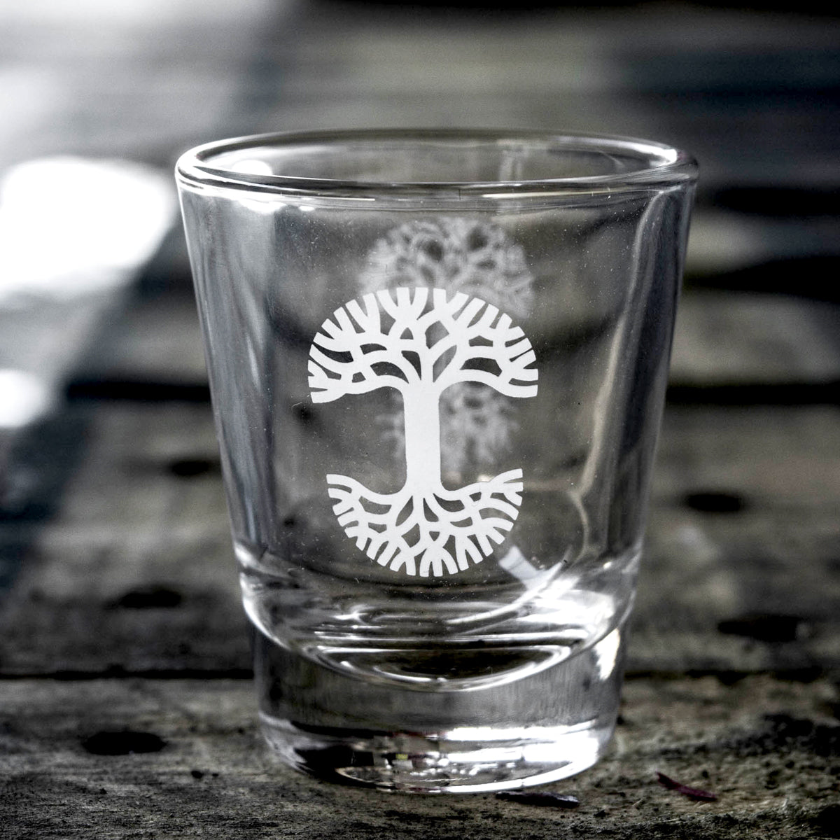 Clear glass liquor shot glass with translucent white Oaklandish tree logo on a picnic table. 