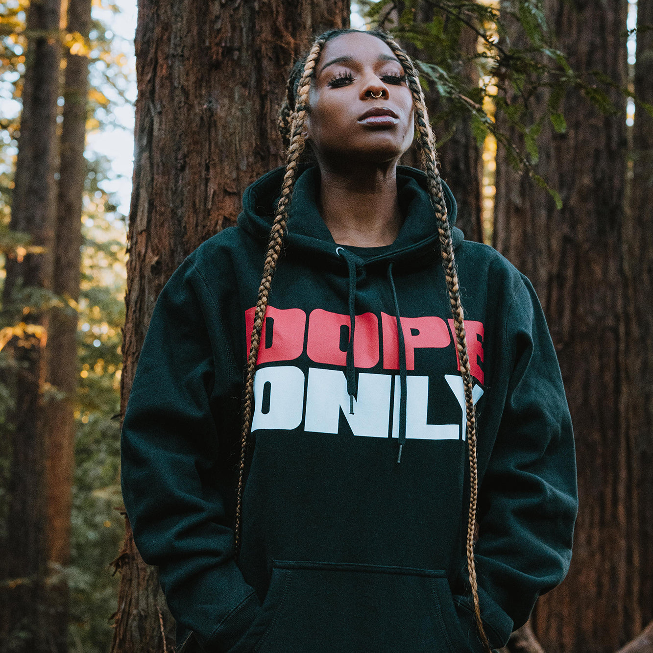 Woman standing in a forest wearing a black hoodie with a large red and white DOPEONLY wordmark logo on the chest.
