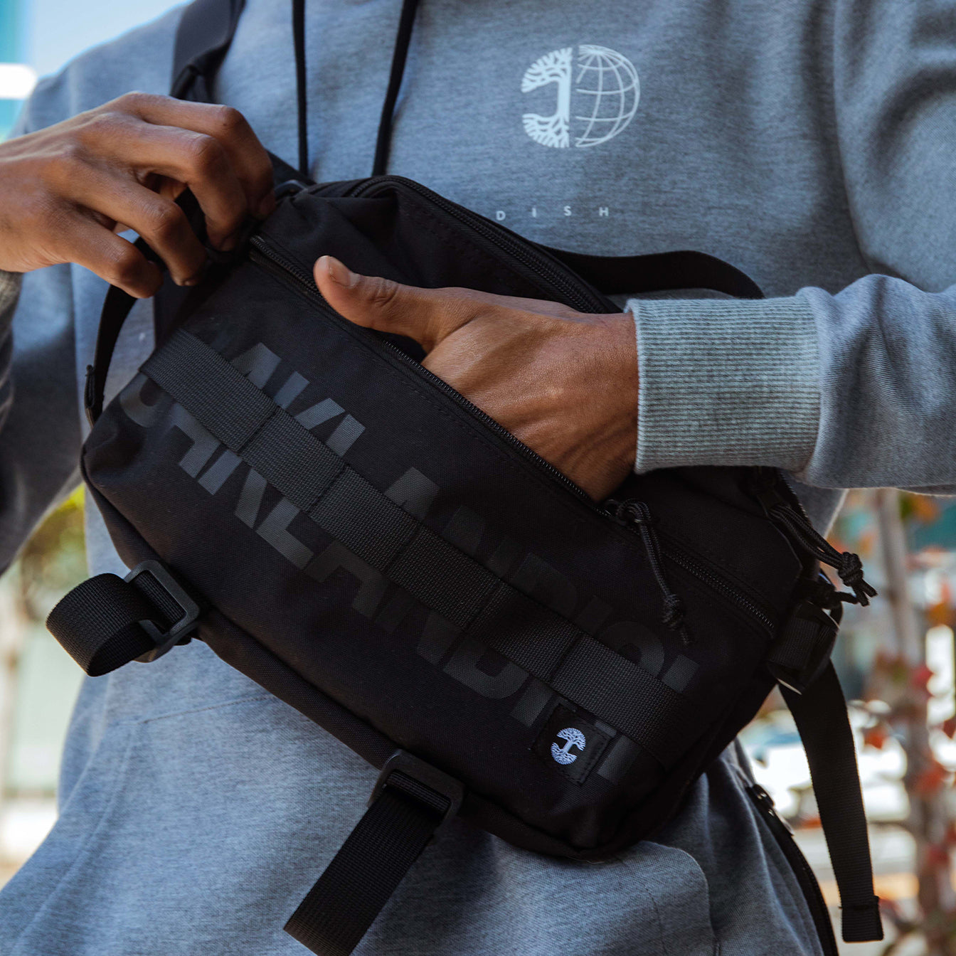 Close-up of the a man’s chest with his hand in a cross body black nylon hip bag with a Oaklandish wordmark and tree logo.