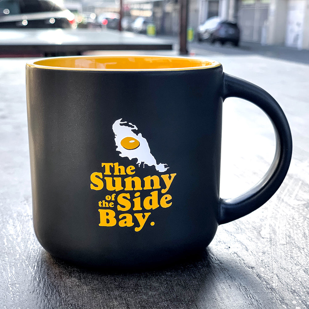 Black mug with yellow inside with picture of Oakland shaped sunny side up fried egg and words The Sunny Side Of The Bay on outdoor bench.