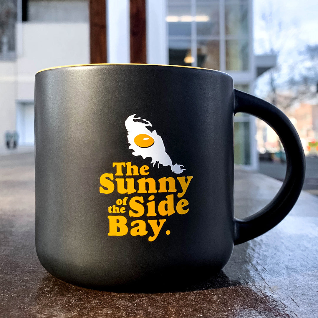 Black mug with yellow inside with picture of Oakland shaped sunny side up fried egg and words The Sunny Side Of The Bay on outdoor bench.