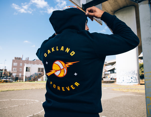 Guy wearing dark navy hoodie with back print with text Oakland Forever around a basketball with a lightning bolt.