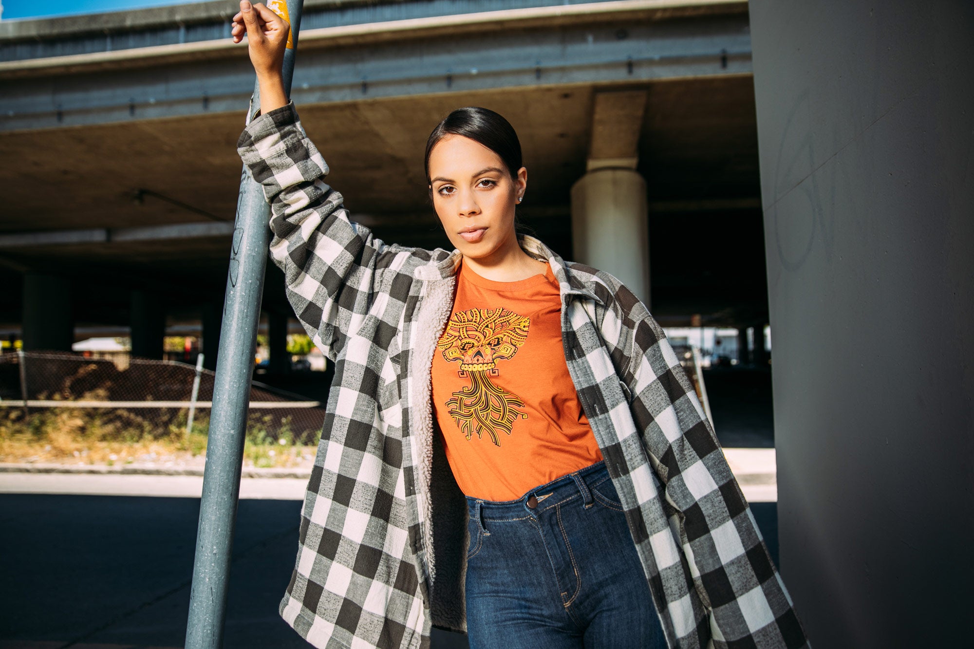 Girl wearing orange tee with plaid shirt over, tee depicts the Ancient Roots design with skull above and tree roots below.