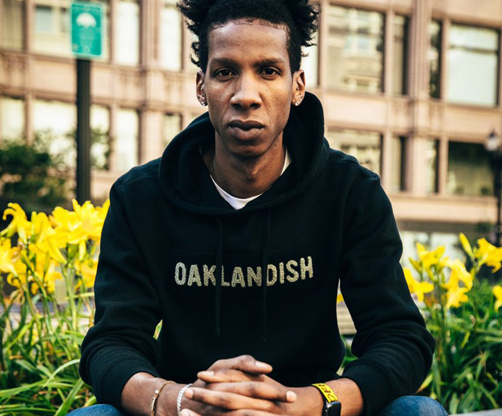 Brandon, seated in front of yellow flowers in Downtown Oakland wearing an Oaklandish hoodie.
