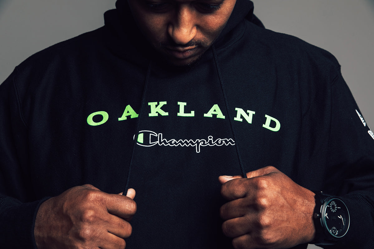 Guy looking down at the front print of his black hoodie, with Oakland in fluorescent green and Champion logo.