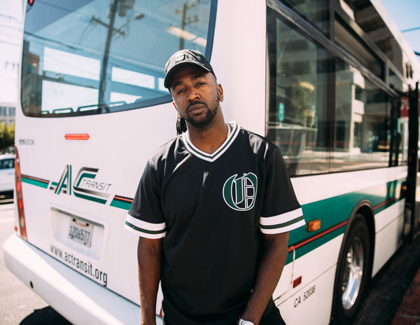 Tunde wearing an Oaklandish jersey in front of an AC Transit bus.