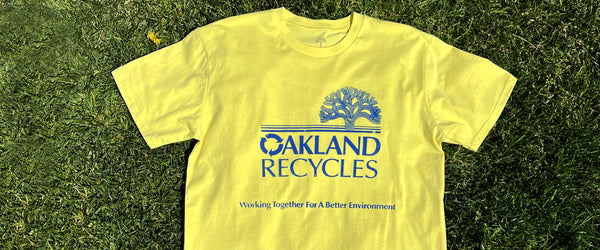 yellow tee laying on grass with blue ink saying Oakland Recycles and blue City of Oakland tree