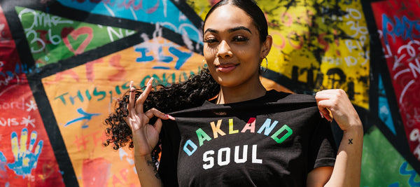 girl with ponytail wearing a black tee with Oakland Soul colorful wordmark in front of graffiti wall