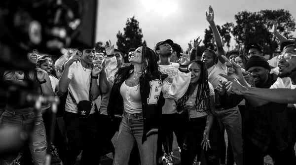 Black and white photo with Goapele singing, wearing varsity jacket and throng of students with her.