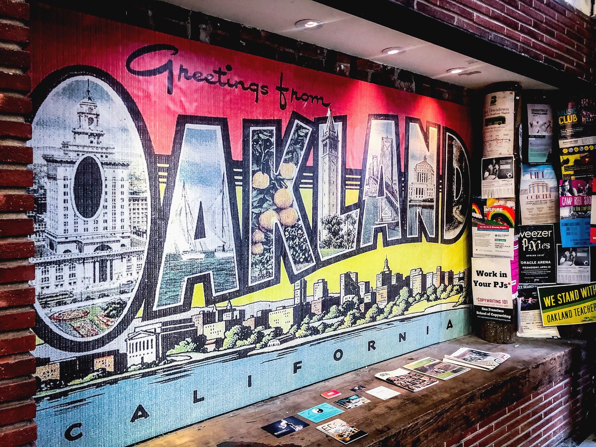 Inset at the flagship store with a vintage postcard style rendering of Greetings from Oakland design featuring iconic imagery inside the letters in OAKLAND and a lakeside skyline in red, yellow, and blue.