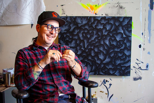 featured artist Jet Martinez, Bay Area Artist & Muralist, sitting with hat on, in front of example of his art