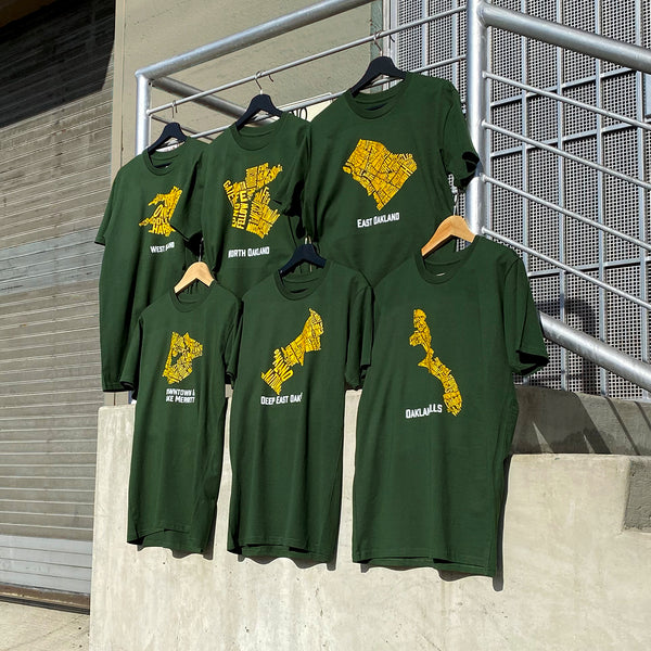 Six green tees from the collection on hangers, hanging from a streetside structure.