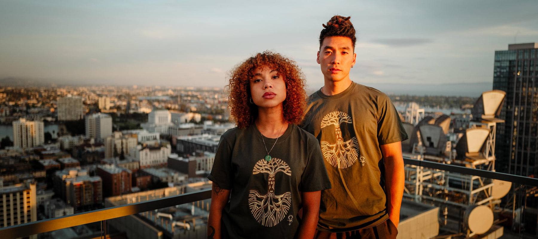 A man and woman on an Oakland rooftop in a cypress green t-shirt with an Oaklandish logo rendered by artist Timothy Bluitt.