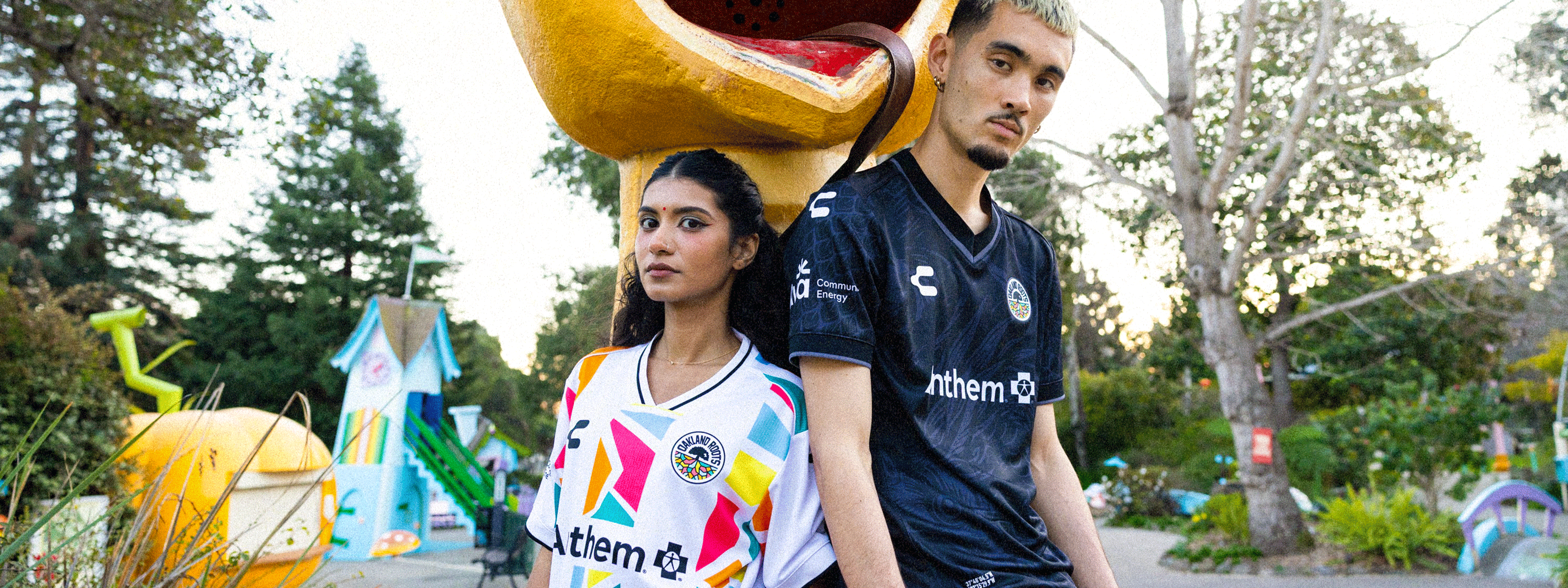 Soccer player in a white and black jersey stands on a sunlit soccer field in an empty stadium, arms behind his back. The sunlight creates a halo effect behind him, highlighting the green grass.