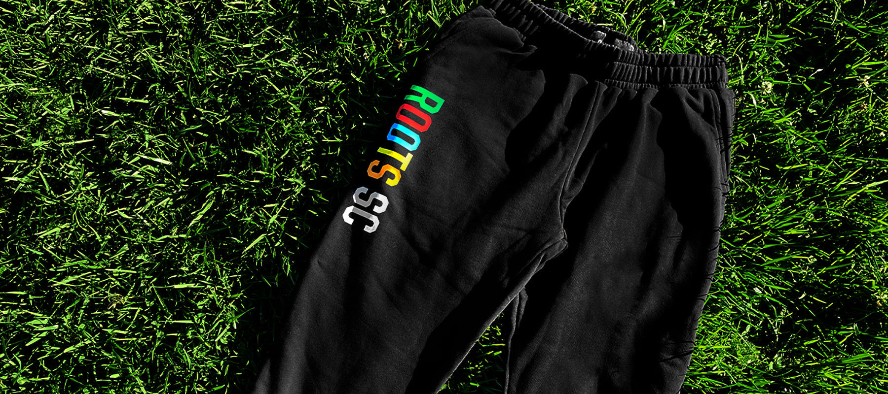 Black Oakland Roots RC joggers with elastic waist and full-color ROOTSSC wordmark on right leg lying outdoors on grass.