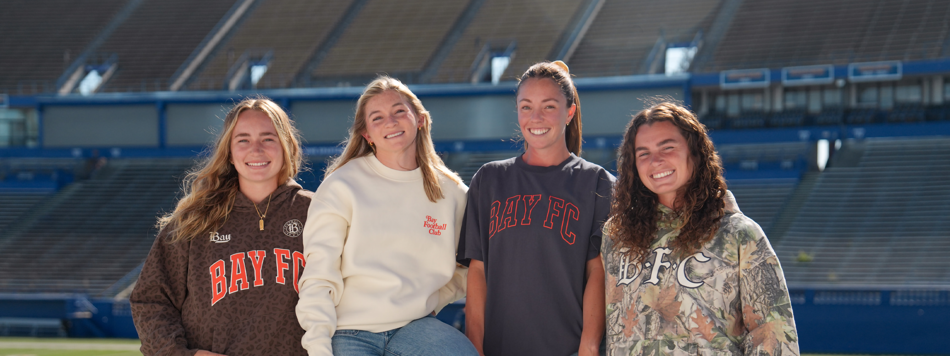 Four women stand together smiling in an empty stadium, wearing Bay FC shirts and sweatshirts in various colors and patterns. The stadium seats are blue and mostly unoccupied.