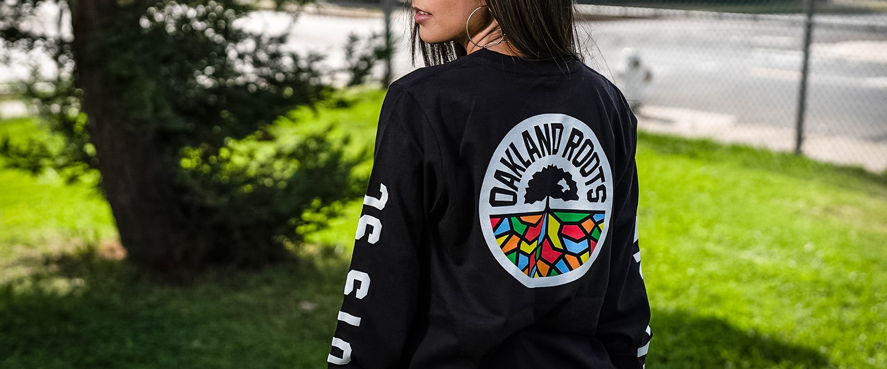 Woman with back to camera head over shoulders, wearing back long sleeve t-shirt with full-color round Roots RC logo graphic.