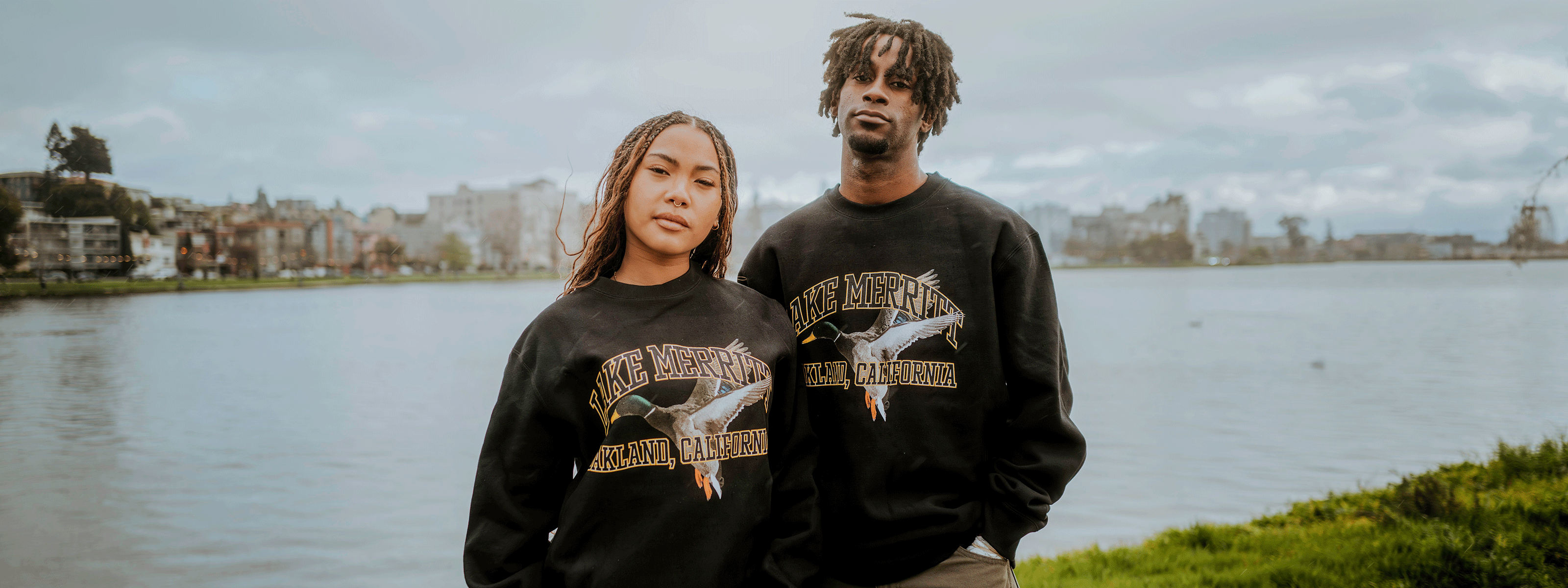 A woman and man stand by a lake, wearing matching "Make America Native Again" sweatshirts. The background features a grassy shore and distant buildings under a cloudy sky.