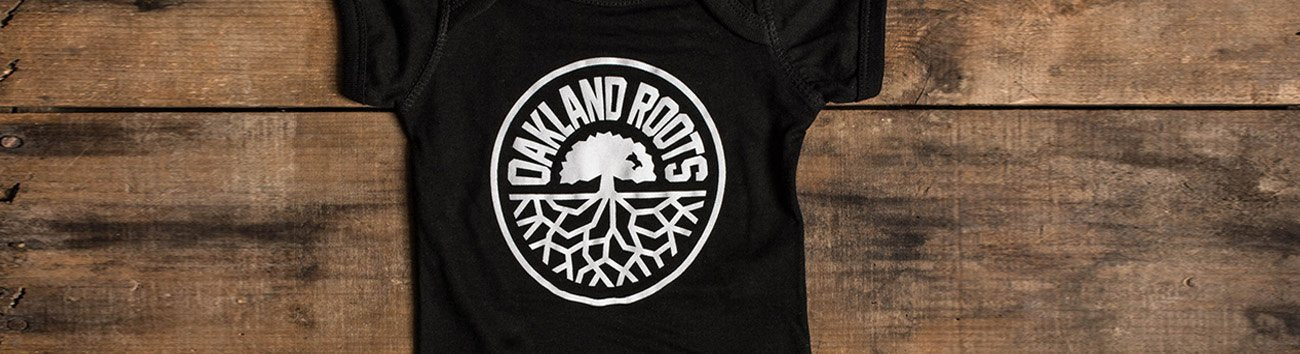 Oakland Roots logo on black one-piece, with wood backdrop.