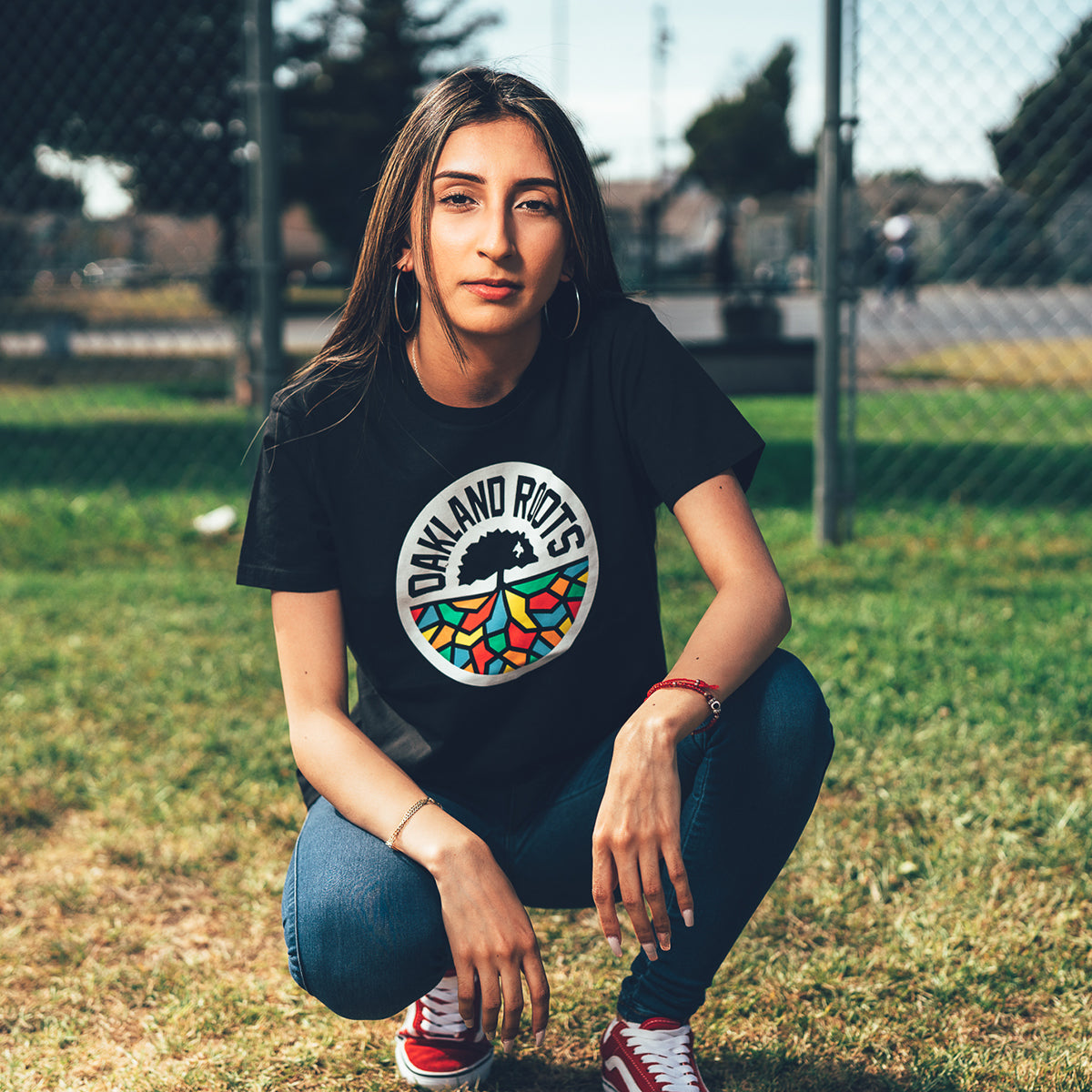 A woman with long brown hair crouches on a grassy field in front of a chain-link fence. She wears the "Women's Oakland Roots SC Classic Tee" by Oakland Roots SC, a black t-shirt made of 100% cotton and featuring the colorful "OAKLAND ROOTS" design. She pairs the tee with blue jeans and red sneakers. Her look is complemented by hoop earrings and a red bracelet. Trees and pavement can be seen in the background.