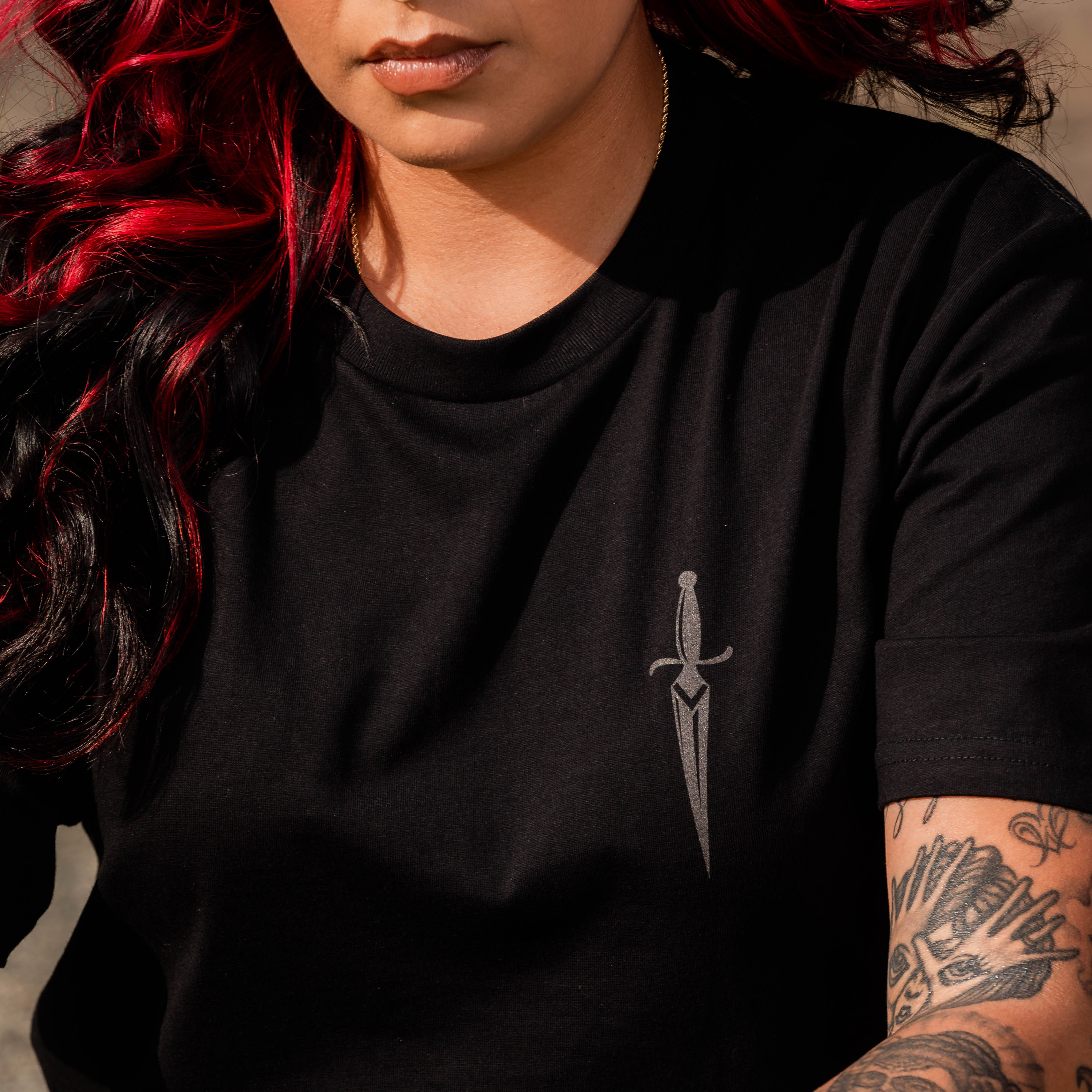 Woman with black and red long hair and tattoos wearing black tee  with dagger front left chest print.