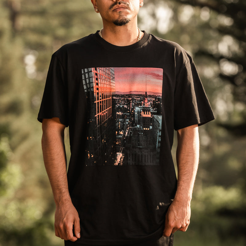 Zoomed in version of male model outdoors facing front with top of head not visible wearing black tee with Vincent James photography image of the Oakland Tribune building and the Downtown Oakland landscape at sunset.