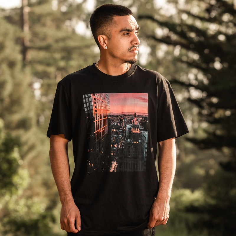 Male model outdoors wearing black tee with Vincent James photography image of the Oakland Tribune building and the Downtown Oakland landscape at sunset.