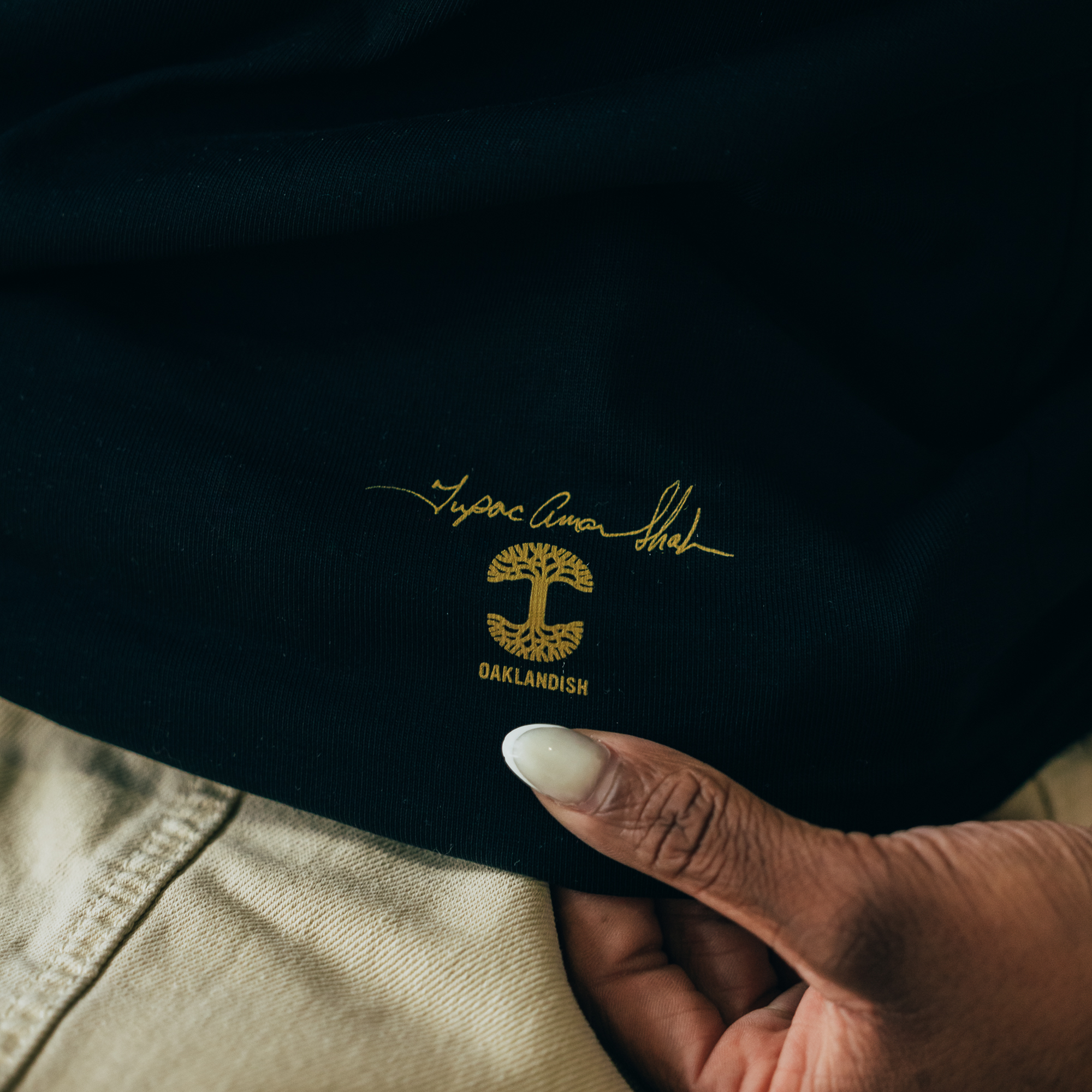 A hand holds the "Against the World Tee" by Oaklandish, showcasing a black fabric with the iconic gold logo of a stylized tree and Tupac-inspired signature. The beige pants in the background convey a casual hip-hop vibe and highlight attention to detail.