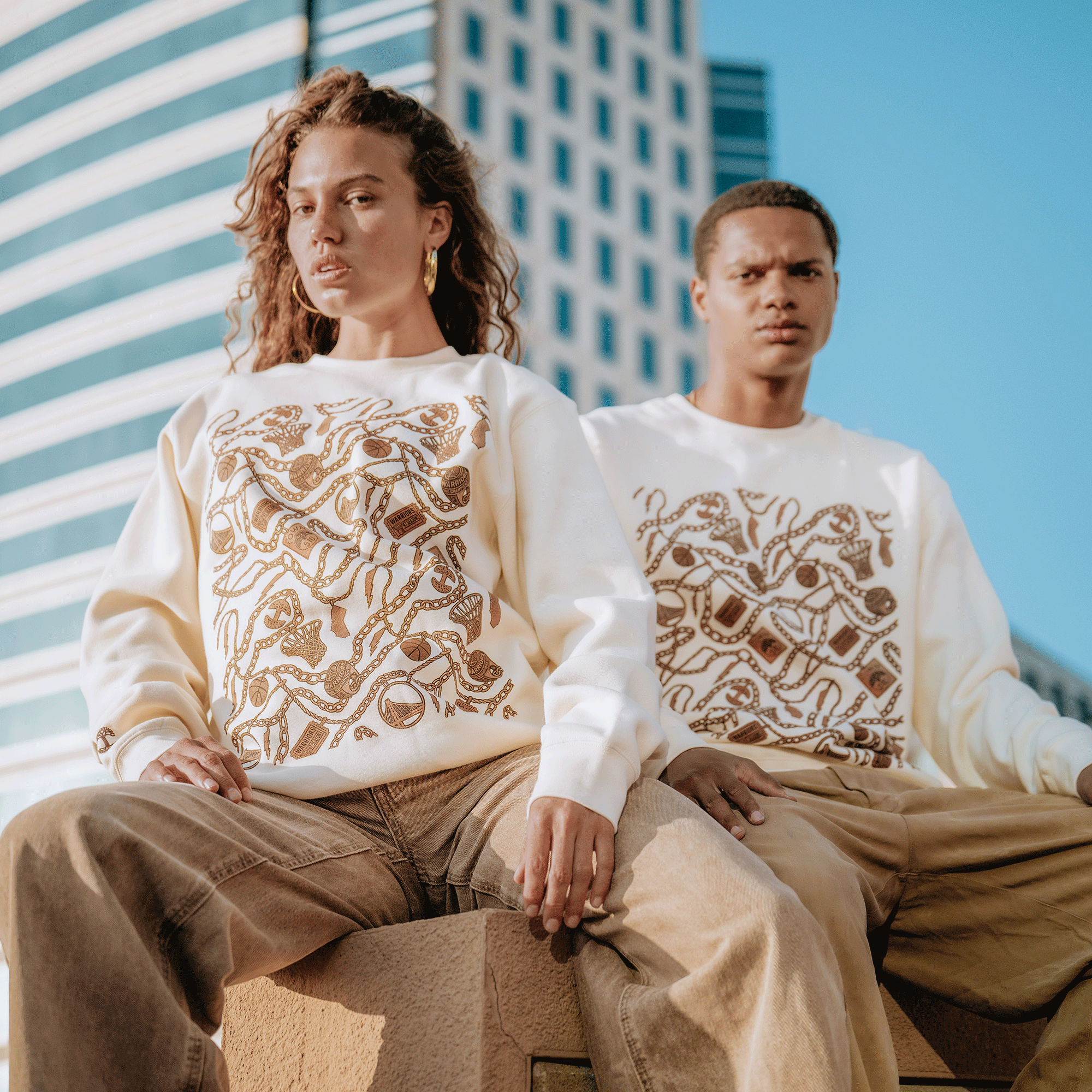 Two people sit on an outdoor ledge in matching GSW All Gold Forever Crew sweatshirts, featuring intricate brown snake designs. Both wear tan pants, with modern glass buildings behind them, channeling a bold Golden State Warriors vibe.