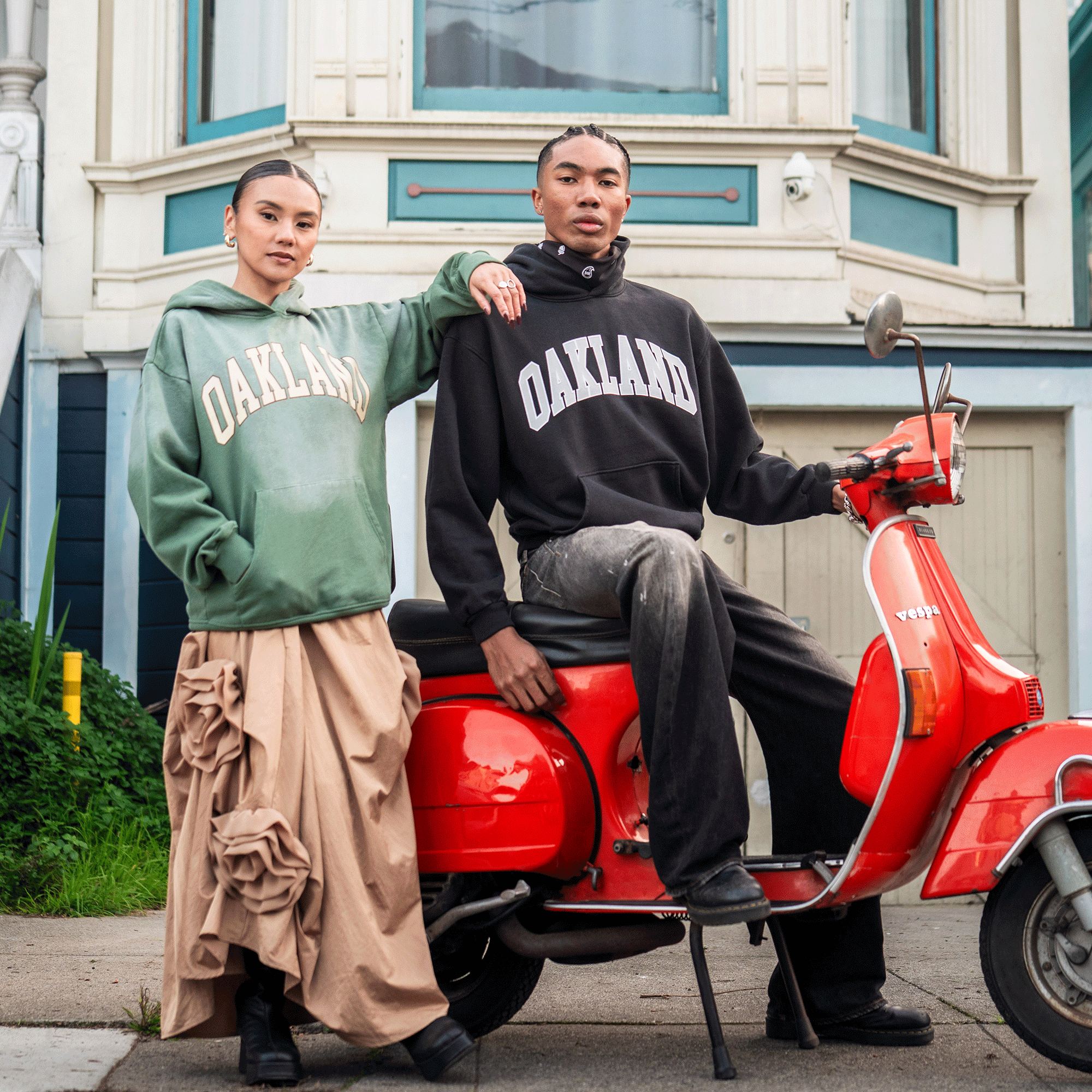 Two people pose with a bright red Vespa on a city sidewalk, both wearing the Oaklandish Alma Mater Hoodie 2.0, made from 100% cotton, with Victorian-style building details in the background.