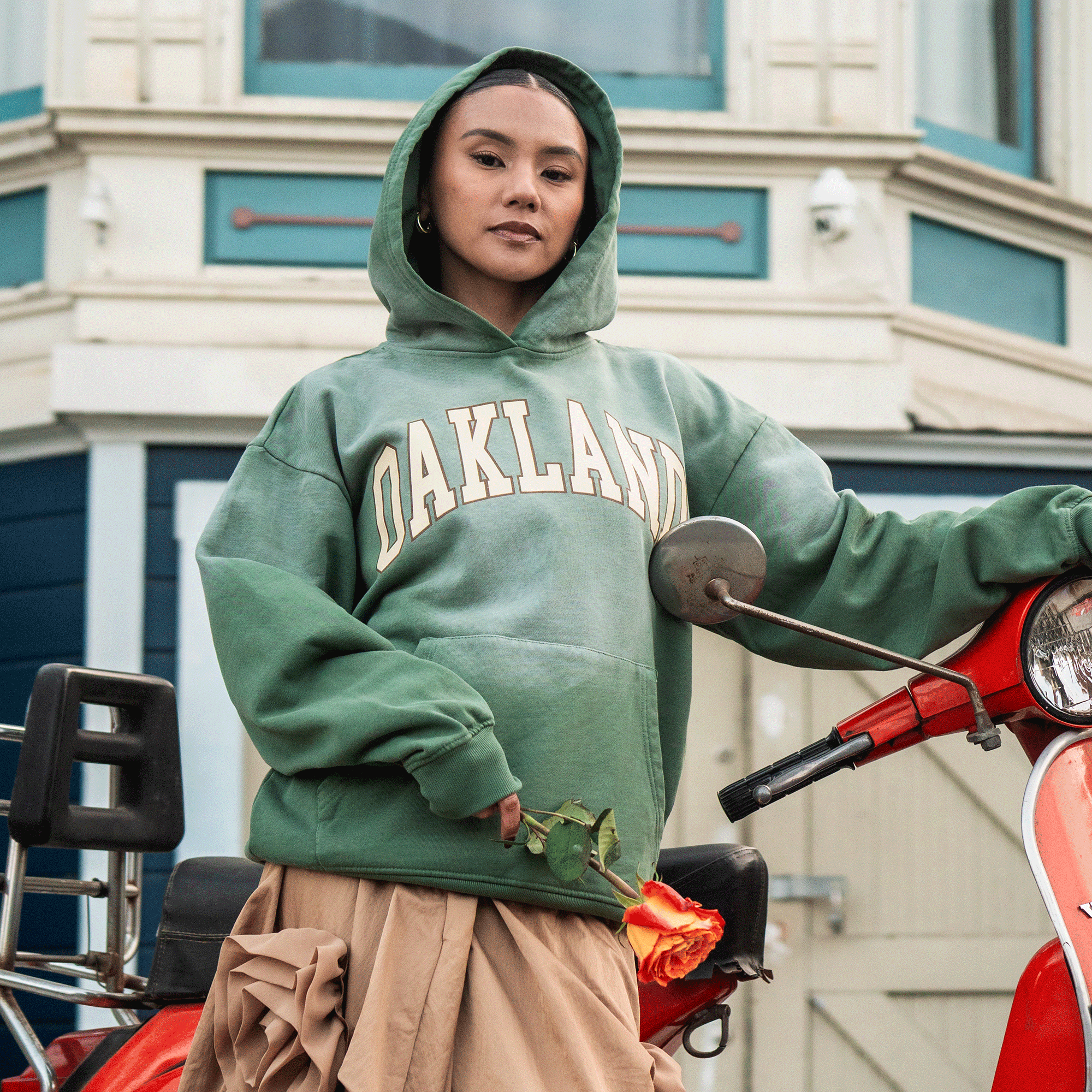 A person in a green Alma Mater Hoodie 2.0 by Oaklandish and a tan skirt leans against a red scooter, sporting hoop earrings and confidence, with blue and beige buildings featuring windows and decorative trim in the background.
