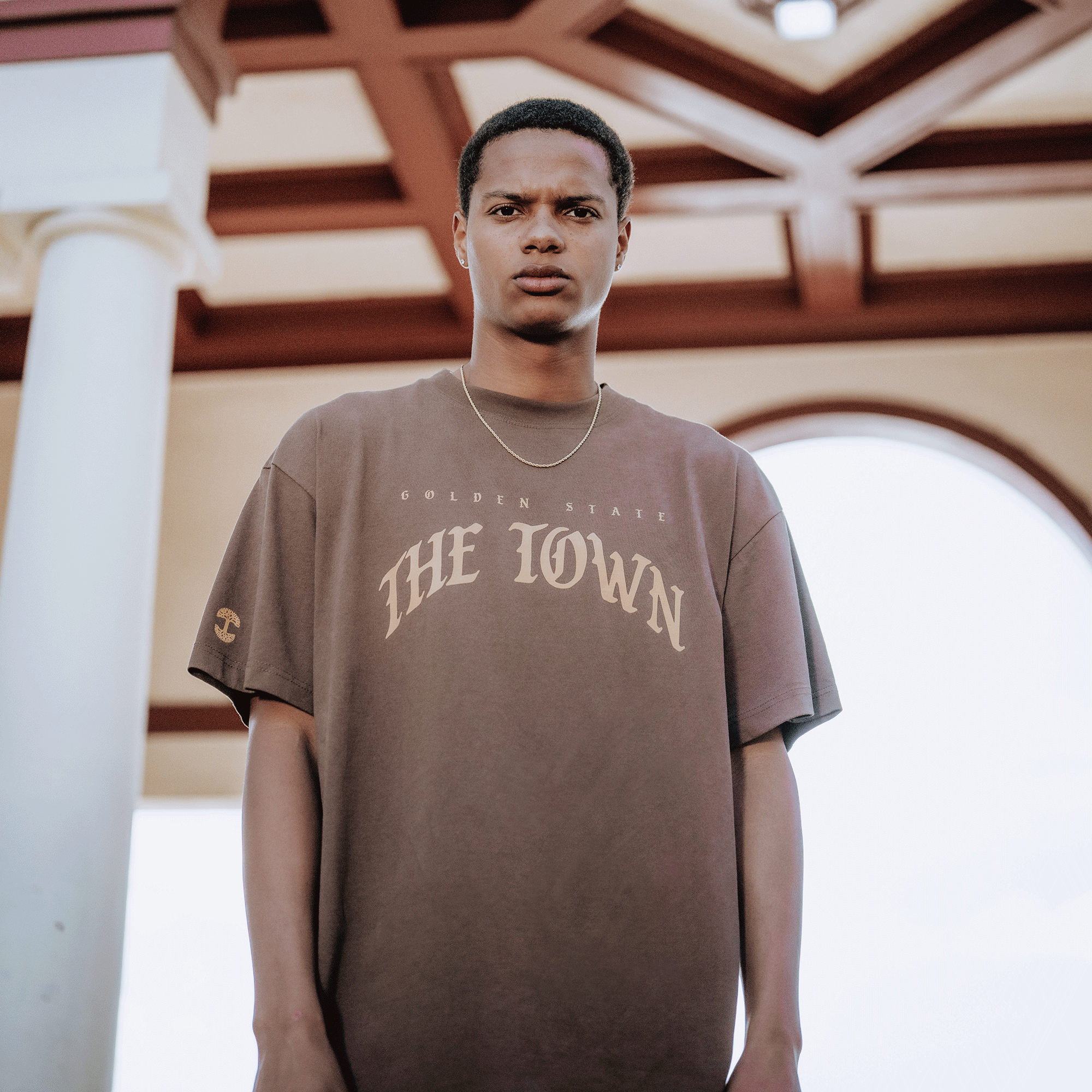 A young man stands outdoors under a decorative ceiling with columns, wearing GSW's Arched Black-Letter Tee. Natural daylight highlights his serious expression—an effortless nod to Golden State Warriors style.