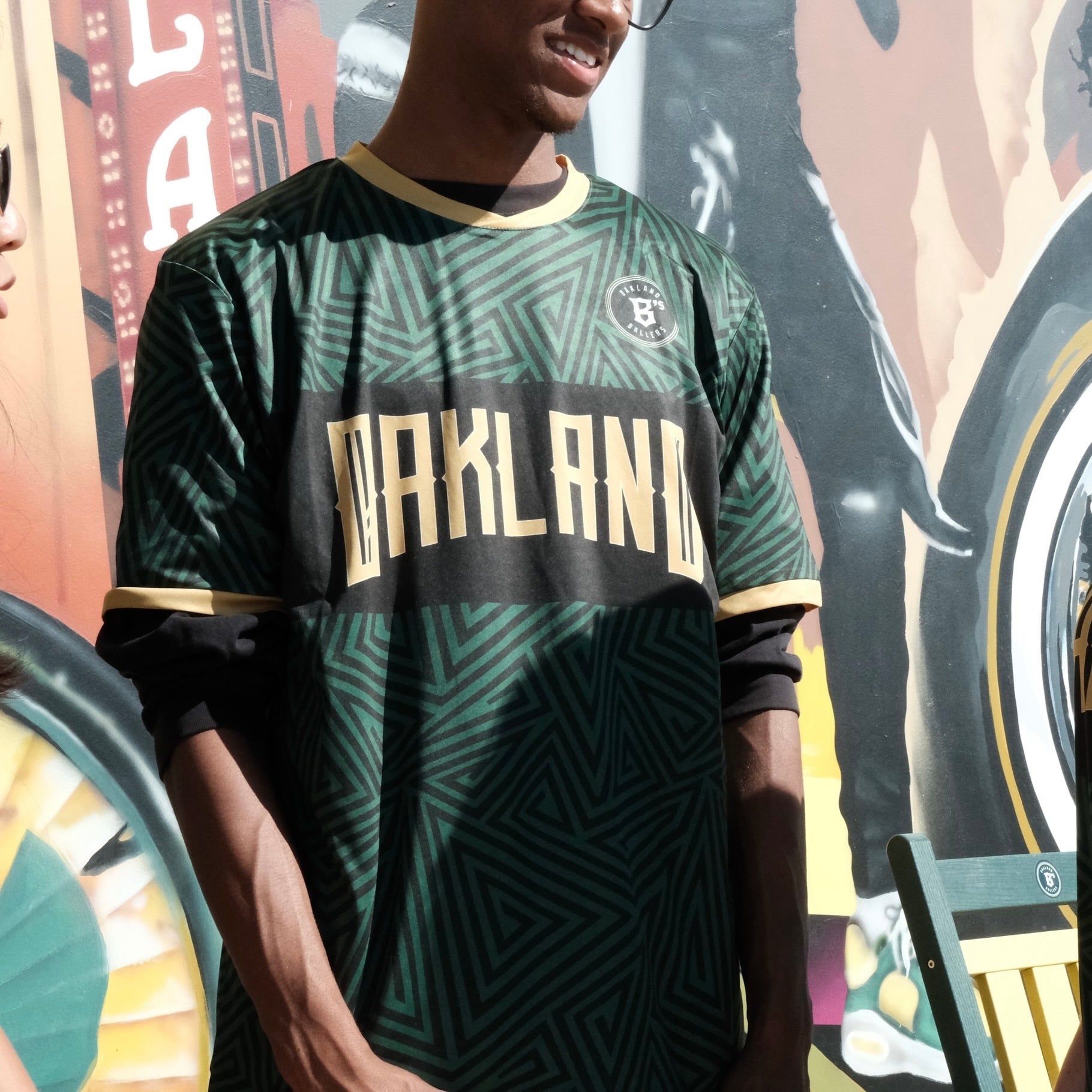 A person wearing glasses and an Oakland Ballers SC Jersey—featuring a green geometric pattern that reflects Oakland Ballers pride—stands in front of a colorful mural, partially visible, with another individual to the left and a green chair to the right. Sunlight casts shadows across the scene.