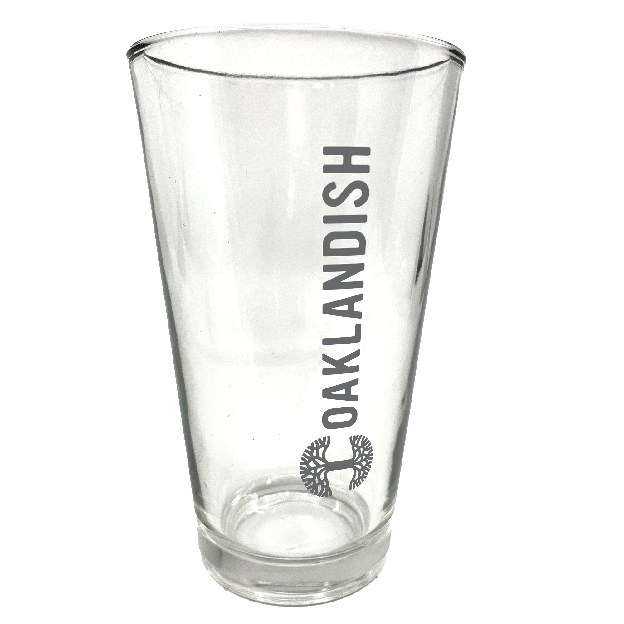 Image of the Oaklandish Banner Pint Glass—a clear, tapered drinkware item featuring the dark grey Oaklandish logo printed vertically. Below the logo is a black image of a spreading oak tree. The background is entirely white, emphasizing the pint glass.