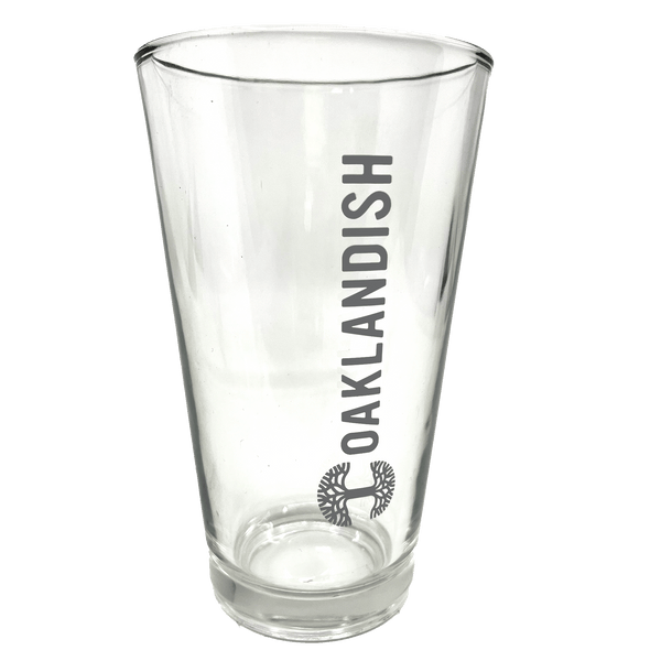 Close-up of clear 16 oz glass beer pint glass with dark grey Oaklandish logo and wordmark imprint.