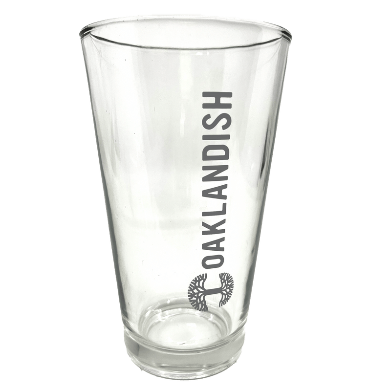 Close-up of clear 16 oz glass beer pint glass with dark grey Oaklandish logo and wordmark imprint.