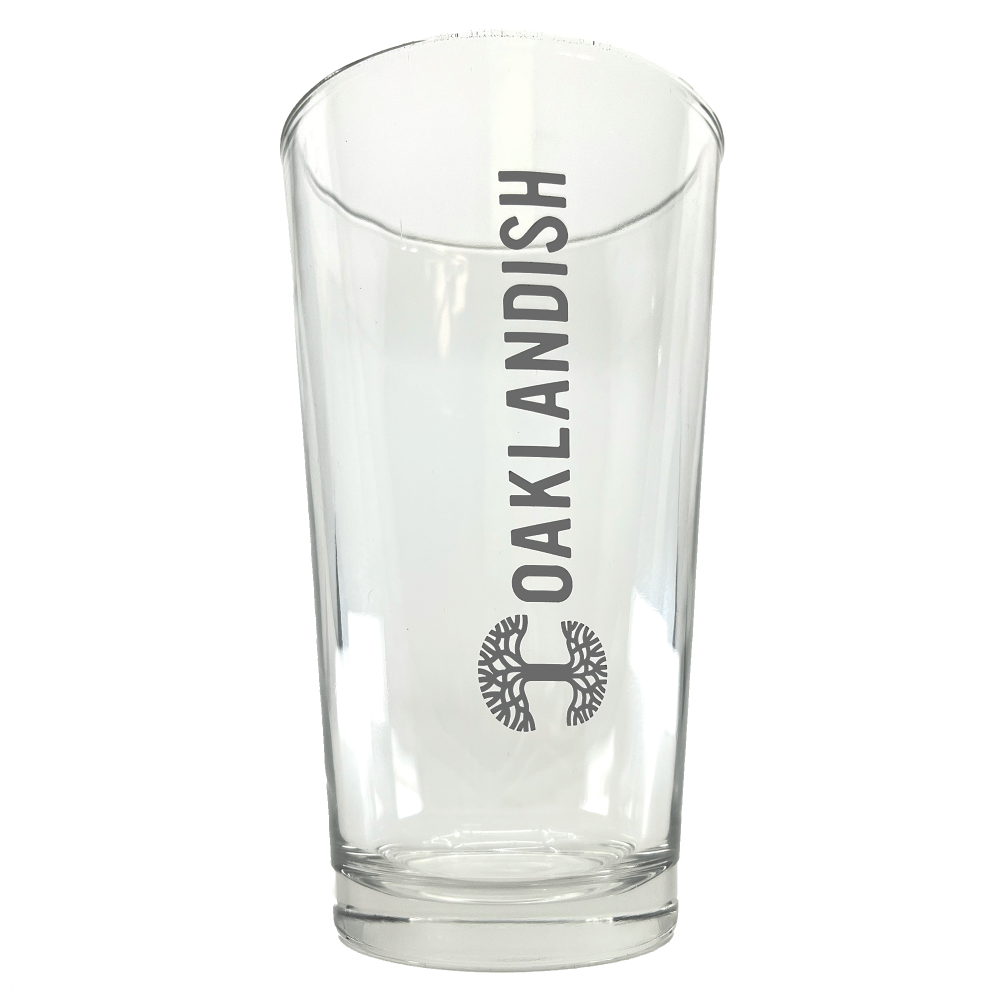 The Banner Pint Glass from Oaklandish is a clear drinking vessel with the word "OAKLANDISH" vertically inscribed in bold, black letters. Underneath this text, you'll find the brand's dark grey logo, a detailed circular tree design. The drinkware has a smooth, slightly tapered shape and features a thick base.