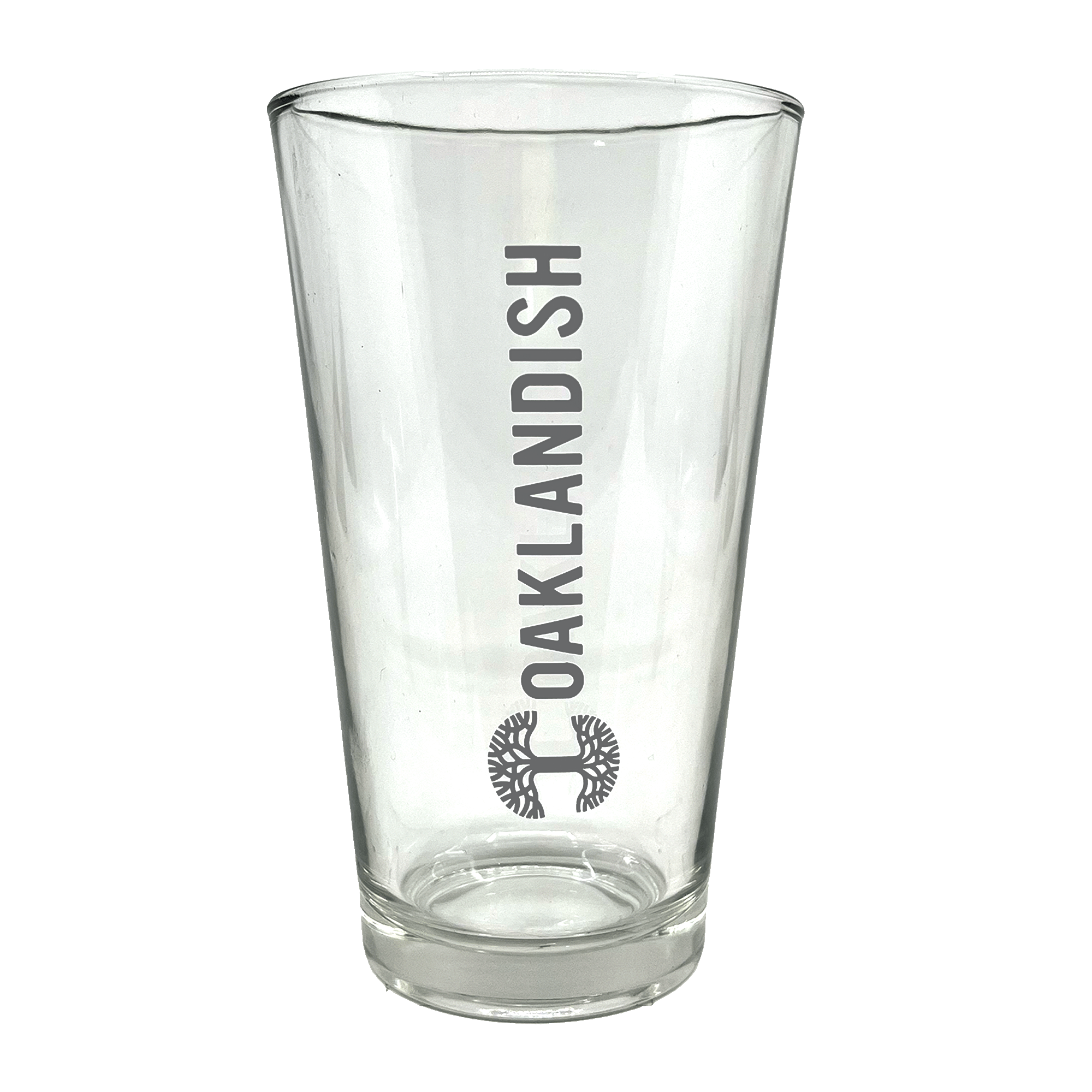 The Banner Pint Glass by Oaklandish is a clear, tall pint glass featuring a bold, dark grey Oaklandish logo in vertical text centered on one side. Below the text is a stylized black tree design within a circular outline. This piece of drinkware boasts a smooth finish and remains empty inside, awaiting your favorite beverage.