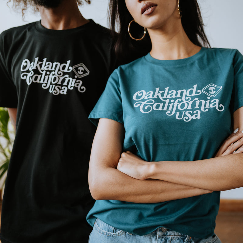 Close up of male model wearing black t-shirt with white Oakland, California USA graphic in cursive and Oaklandish logo and female model wearing atlantic bookman tee.