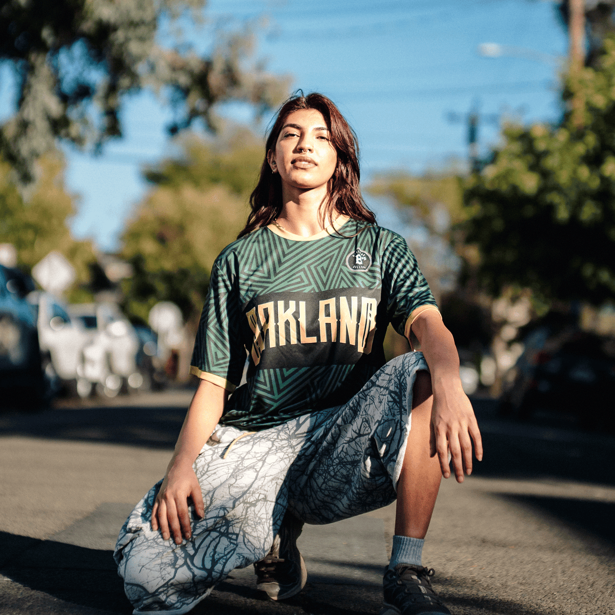 A person with long brown hair, wearing a green and gold Oakland Ballers SC Jersey, poses outdoors on a sunlit street while kneeling on the pavement in light-colored patterned shorts and athletic shoes.