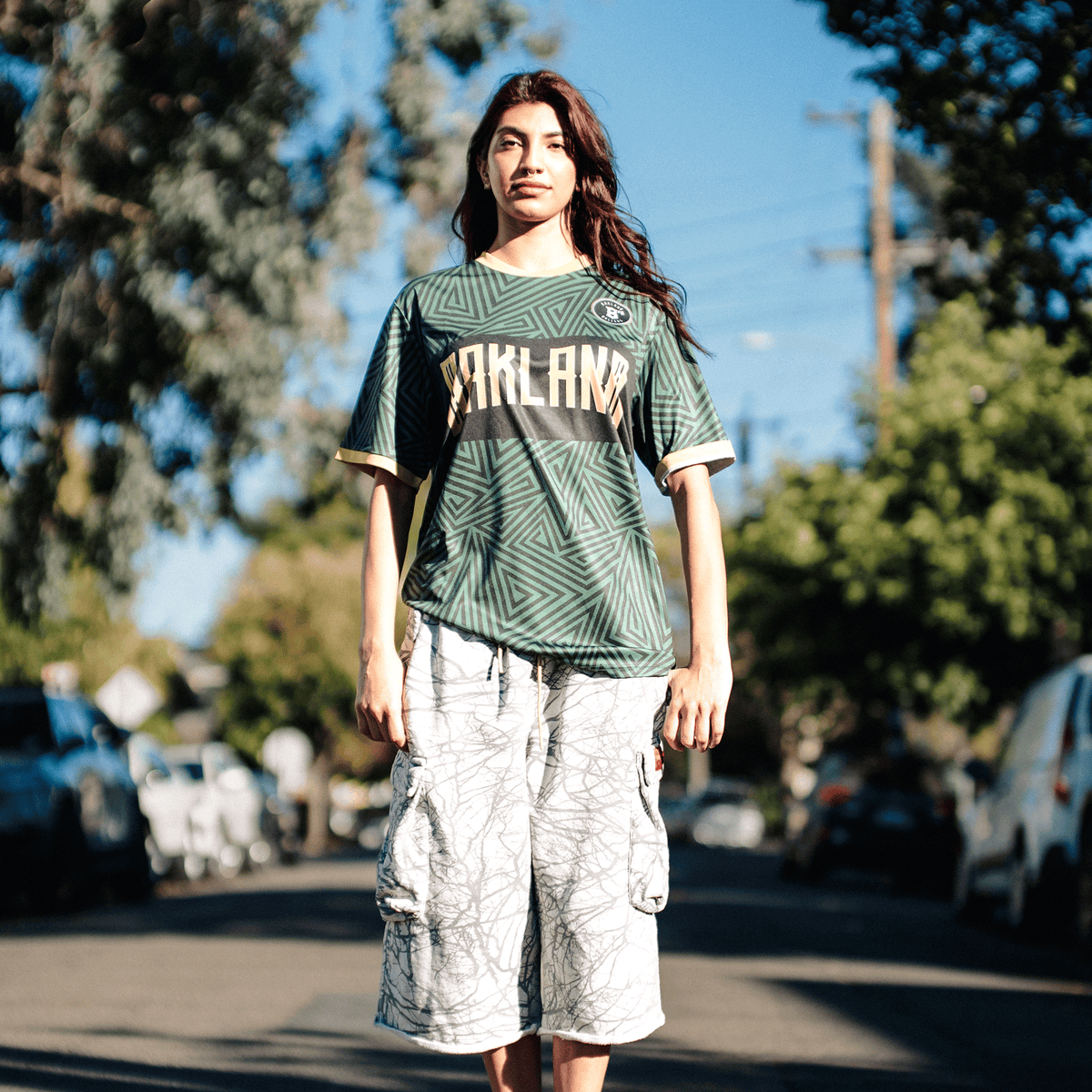 A person with long dark hair stands on a sunlit residential street wearing an Oakland Ballers SC Jersey, which is green with a gold horizontal chest stripe and a geometric pattern, paired with white, knee-length cargo shorts featuring a dark, tree-branch-like print.