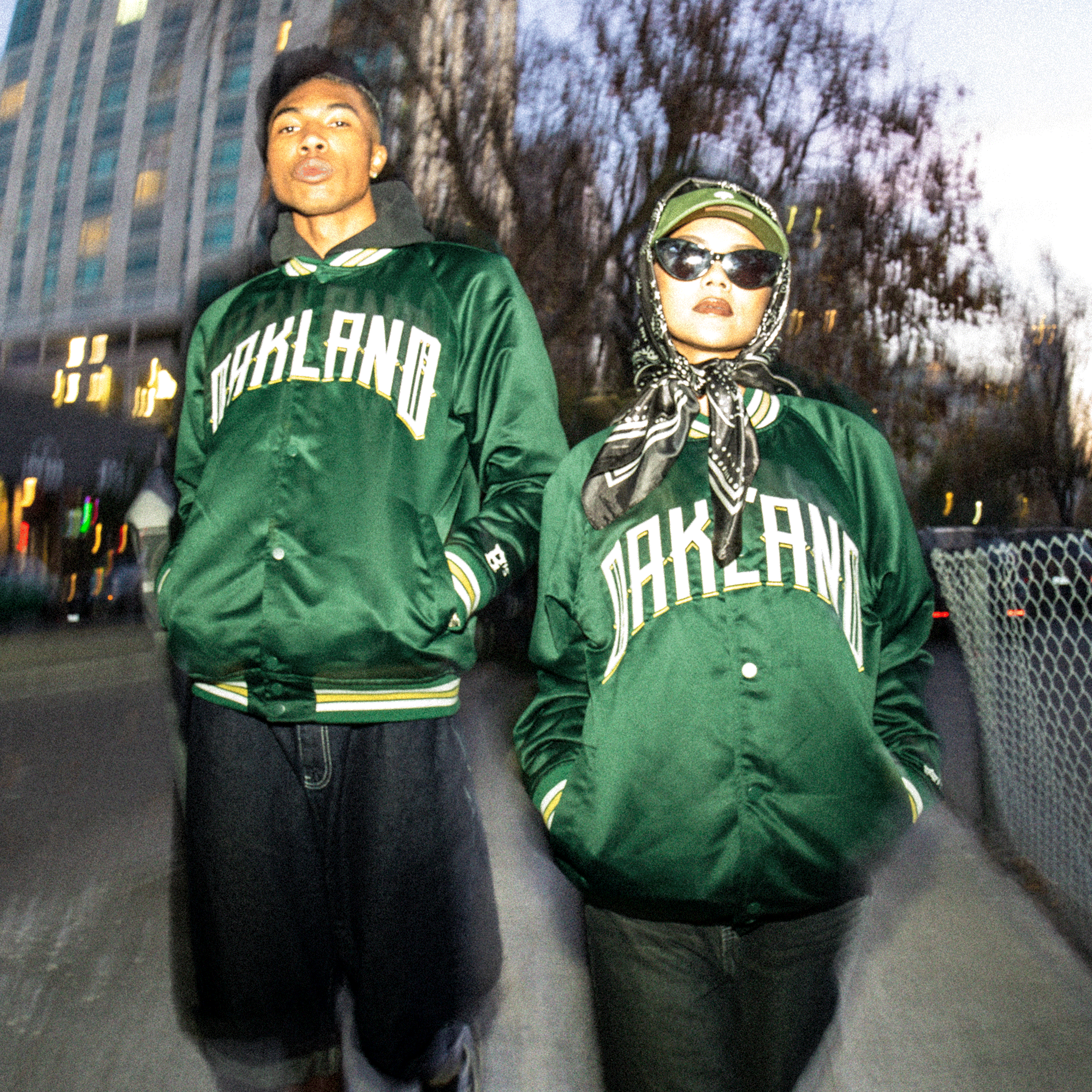 Two people wear matching Oakland Ballers Satin Jackets outdoors—one with short hair and dark pants, the other with sunglasses and a scarf. The blurred evening backdrop features tall buildings and trees.