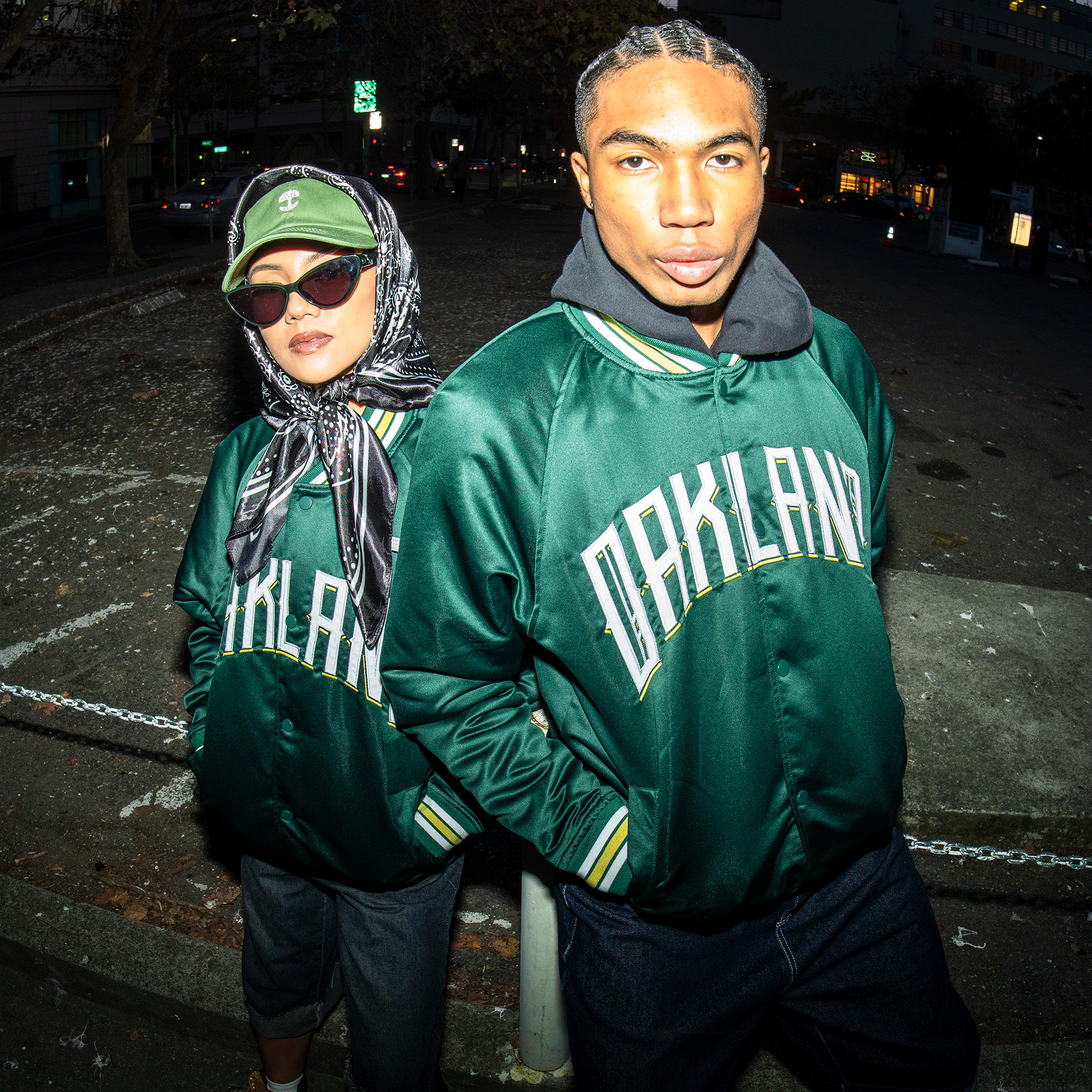 Two people pose confidently outdoors at night, both wearing the Oakland Ballers Satin Jacket by Oakland Ballers. One pairs it with sunglasses, a patterned scarf, and cap; the other styles cornrow braids and a hoodie under the jacket.