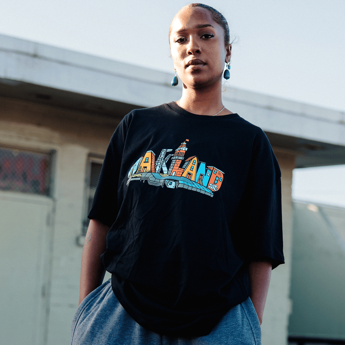 A person with hair pulled back wears a black Oaklandish Blockletters Tee featuring a stylized Oakland skyline graphic, grey sweatpants, and dangling blue earrings while standing outdoors against a plain, light-colored building.