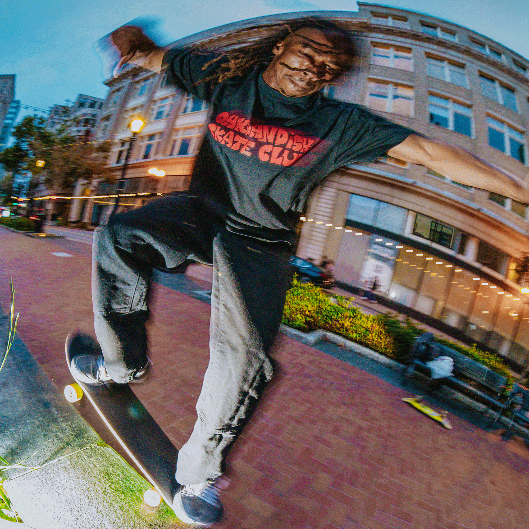 A skateboarder with dreadlocks, wearing the Oaklandish Skate Club Tee and black pants, grinds a ledge at dusk in the city. Captured with a fisheye lens, the shot highlights skateboard culture against an urban backdrop.