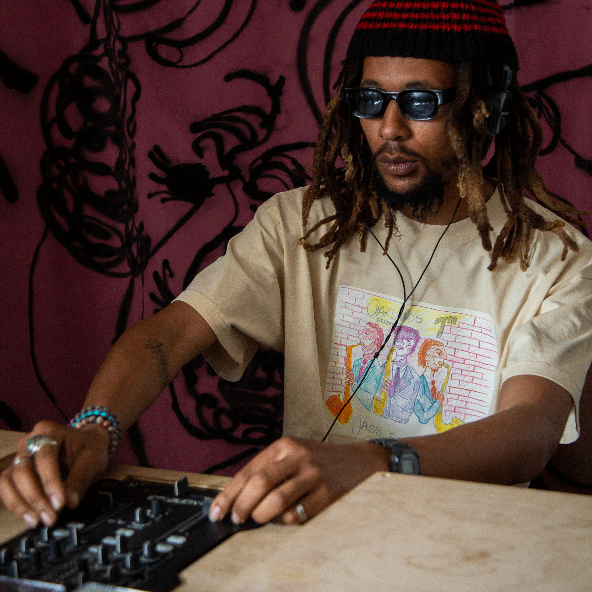 A man with dreadlocks and sunglasses wears the Oaklandish 7th St. Players Tee—100% cotton, classic fit with a colorful cartoon graphic—plus headphones, bracelets, a red-striped beanie, and works a DJ mixer before a pink wall with abstract designs.